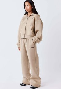 Sudadera con capucha de forro polar beige con cremallera frontal y grandes bolsillos, combinada con pantalones anchos a juego. Ambos presentan sutiles detalles con el logo.