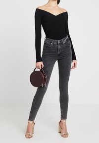 Body noir côtelé à épaules dénudées associé à un jean skinny gris et des sandales beiges à talons blocs, tenant un sac rond bordeaux.