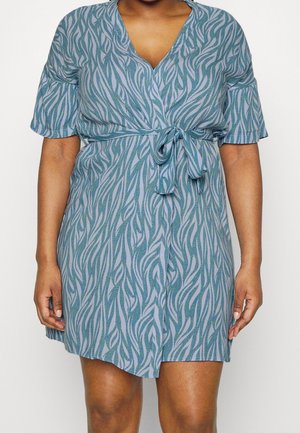 Woman wearing short-sleeve blue dress with green and white wavy stripes and a tied belt at the waist.