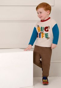 Camisa de manga larga beige con el colorido texto "VIDA DE BICHOS", combinada con pantalones marrones y zapatillas multicolores. Superficie suave, diseño casual.