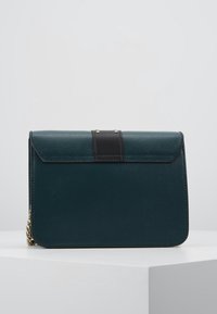 Clutch in pelle texture verde scuro con dettaglio della tracolla nera e una catena dorata parzialmente visibile sul lato sinistro, poggiato su una superficie bianca.