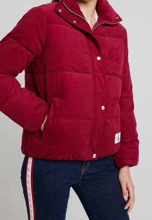 Woman wearing a red padded jacket with snap buttons and side pockets, paired with dark jeans featuring a branded side stripe.