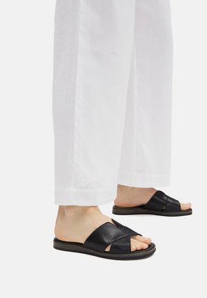 Čierne sandále s krížovými remienkami vyrobené z textúrovaného materiálu, s plochou podrážkou. Nosené s voľnými bielymi skrátenými nohavicami.