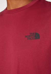 Mörkröd bomullströja med en liten svart logotyp på vänster bröstområde. Tyget har en mjuk textur och en klassisk rundad halslinning.