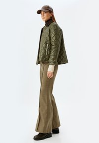 Olivgrüne Steppjacke, brauner Kragen, cremefarbener Rollkragenpullover, karierten Schlaghosen, braune Stiefel und eine braune Mütze. Strukturierte und texturierte Details.