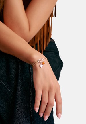 Hand einer Frau, die auf dunklem Denim ruht, trägt ein beiges Armband mit Herz- und Bernsteinanhängern, im Hintergrund ist ein braunes Fransenoberteil sichtbar.