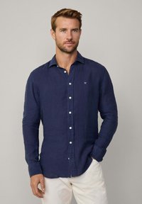 Navy blue linen shirt with a button-down collar, front placket with white buttons, long sleeves, and embroidered logo on the chest.