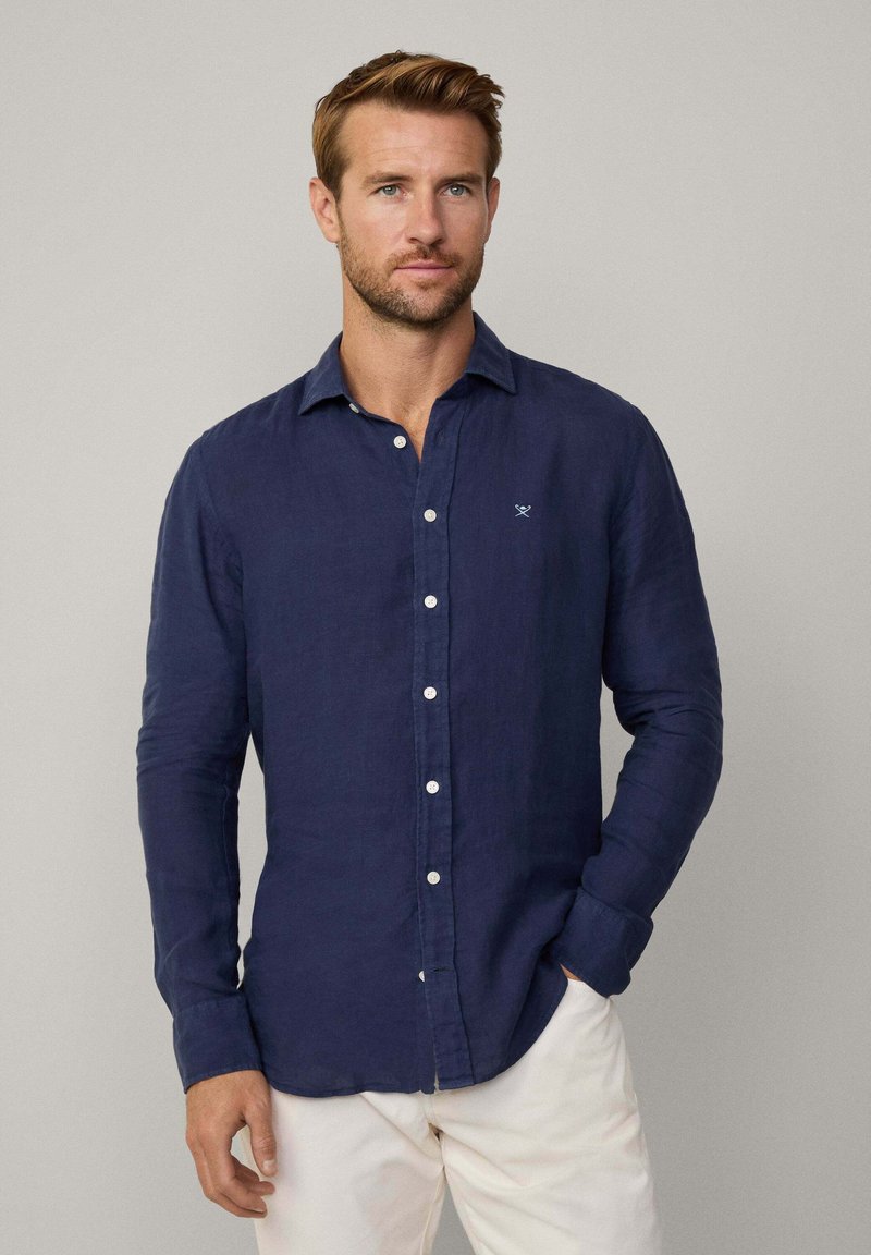 Camisa de lino azul marino con cuello botón, tapeta frontal con botones blancos, mangas largas y logotipo bordado en el pecho.