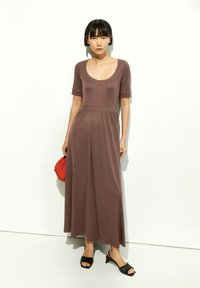 Brown scoop-neck maxi dress with short sleeves, fitted bodice, flowing skirt. Worn with black slip-on shoes and a red bag. Smooth texture.