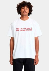 Hombre con cabello rizado que lleva una camiseta blanca con texto rojo que dice: "Cuando muera, voy a atormentarte hasta el infierno."