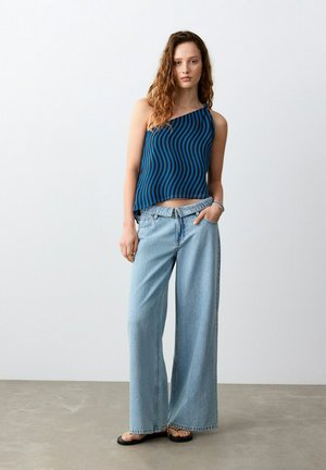 Femeie purtând un top albastru și negru, cu o singură bandă pe umăr și dungi ondulate, blugi largi, de culoare albastru deschis, și sandale negre, stând în fața unui fundal simplu.