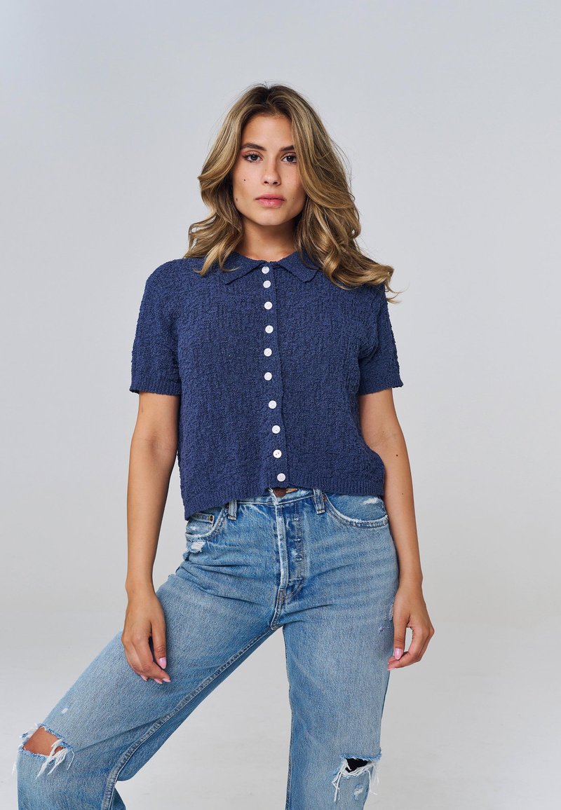 Femme aux cheveux blonds ondulés portant un haut bleu marine à manches courtes boutonné et un jean bleu clair déchiré, debout avec une jambe légèrement levée.