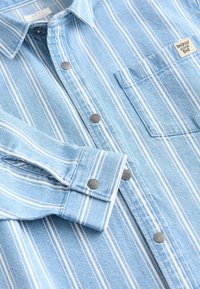 Chemise bleu clair à rayures verticales blanches, boutons-pression en métal, poche poitrine avec étiquette « happy cool kid » et poignets boutonnés.