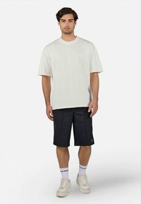 Tricou scurt de culoare bej deschis, purtat cu pantaloni scurți bleumarin, având o textură fină. Accesoriile includ șosete albe și adidași crem.
