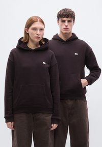 Σκούρες καφέ μπλούζες με κουκούλα, εμπρός τσέπη και ριμπ μανσέτες, με μικρό κεντημένο λογότυπο στο στήθος. Φοριούνται από δύο μοντέλα.