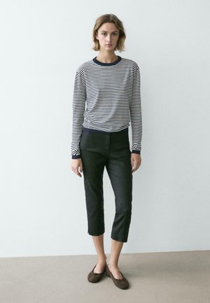 Junge Frau steht vor einer schlichten weißen Wand, trägt ein gestreiftes Langarmshirt, schwarze, knöchellange Hose und braune Ballerinas.