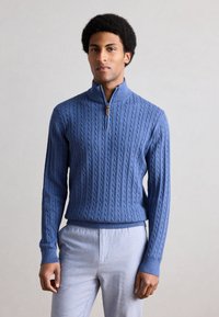 Μπλε μάλλινο πουλόβερ με γιακά τεταρτημόριου φερμουάρ, με σχέδιο πλεκτού καλωδίου, ριμπ μανσέτες και τελείωμα, σε συνδυασμό με ανοιχτόχρωμο παντελόνι.