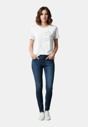 Donna in piedi con le mani nelle tasche, indossa una maglietta bianca con disegni di stelle, jeans skinny blu e sneakers bianche su uno sfondo semplice.