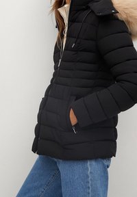 Zwarte pufferjack met een rits aan de voorkant, doorgestikte ontwerp en een met bont gevoerde kap. Voorzien van twee zijzakken en een contrasterende binnenvoering.