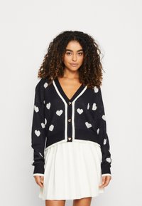 Marine gebreide cardigan met witte hartpatronen, V-hals en crème afwerking. Bevat gouden knopen, een relaxte pasvorm en lange mouwen.