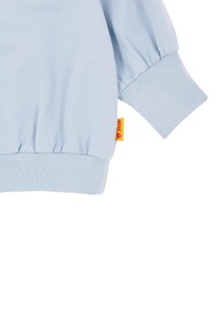 Hellblaues Langarmshirt aus weichem Material, mit schoppenärmeln und einem gerippten Bündchen. Enthält ein kleines gelbes Etikett mit einem Bärenlogo.