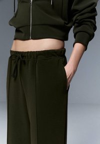 Dunkelgrüner, kurzer Hoodie mit Reißverschluss und Jogginghose mit elastischem Bund und Seitentaschen. Glatte Stoffstruktur.