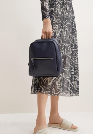 Sac à dos en cuir marine avec finition texturée, poche avant zippée, forme arrondie et poignées supérieures, porté par une personne en robe à motifs.