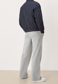 Personne portant une veste bomber noire et un pantalon de survêtement gris ample avec des baskets blanches, debout les mains dans les poches, face à l'opposé.