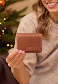 Frau hält eine braune Leder-Geldbörse von Gusti Leder vor einem festlich geschmückten Weihnachtsbaum und trägt einen beigen Pullover.