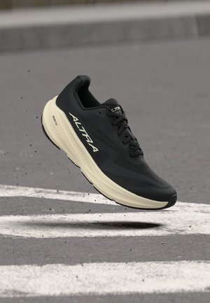 Black and beige Altra running shoe with "Altra" logo mid-air above asphalt road with white markings and small debris.