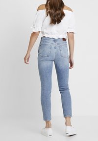 Jeans ajustados de tiro alto en azul claro, con un ligero desgaste y detalles desgastados en los bolsillos traseros, combinados con una blusa blanca de hombros descubiertos.