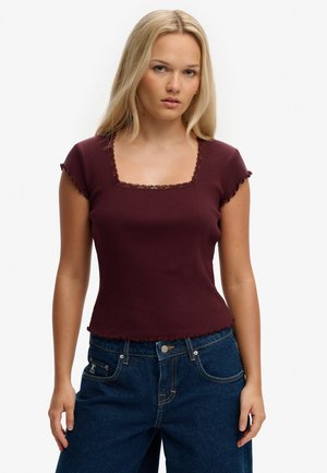 Lyshåret kvinde iført en tætsiddende, kortærmet, burgunderrød ribstrikket top med blonder og højtaljede mørkeblå jeans mod en hvid baggrund.