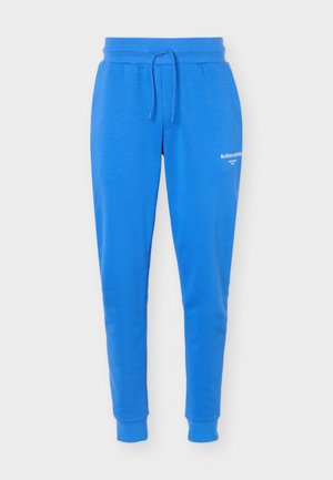 Pantaloni della tuta blu con gamba affusolata, vita elasticizzata e coulisse, polsini alle caviglie e logo testuale bianco sulla parte superiore della coscia sinistra.