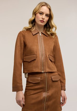 Mujer con cabello rubio ondulado, vistiendo una chaqueta marrón con cremallera y falda a juego con bolsillos de solapa, sobre un fondo neutral.