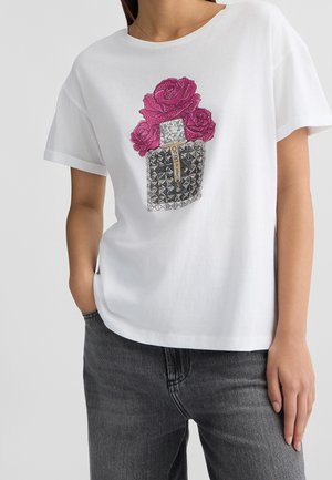 Frau trägt ein weißes T-Shirt mit Grafik einer pinken Rose und eines mit Nieten verzierten Parfümflakons, kombiniert mit verblassten schwarzen Jeans, steht mit einer Hand in der Tasche.