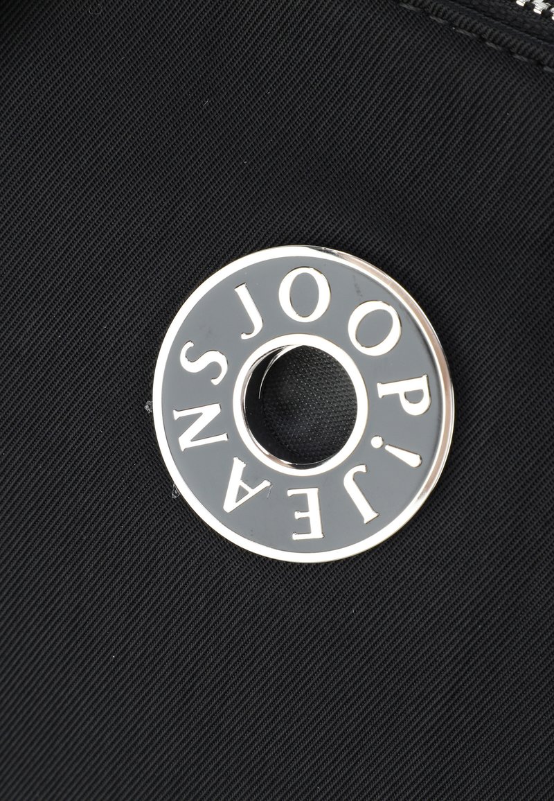 Tejido de nailon negro con un acabado metálico redondo y brillante en plata que presenta el texto "JOOP! JEANS" alrededor de una abertura circular.
