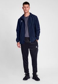 Marineblå vindtett jakke med tettsittende design, kombinert med svarte smale treningsbukser med sidestriper, brukt med svarte sportssko.