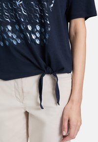 Mørkeblå t-shirt med knudet frontdesign, lavet af blødt stof med et tekstureret mønster trykt i metalliske nuancer. Lysebeige bukser.