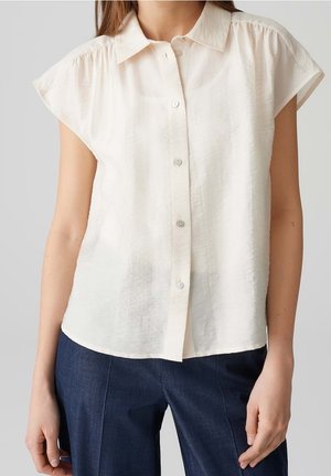 Kvinde iført en cremefarvet skjorte med korte ærmer og knapper, kombineret med mørkeblå højttaljede bukser, stående foran en neutral baggrund.