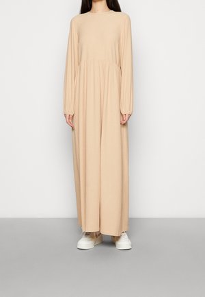 Kobieta w beżowej sukience z długim rękawem sięgającej do kostek, zestawionej z białymi trampkami, stojąca na tle jednolitego białego tła.