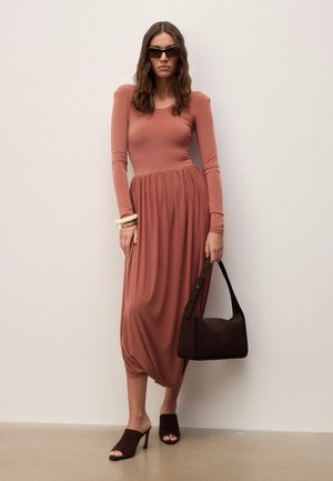 Nainen, joka on pukeutunut pitkähihnaiseen ruosteenväriseen mekkoon, tummiin aurinkolaseihin ja ruskeisiin korkokenkiin, pidellen mustaa olkalaukkua ja seisomassa valkoista seinää vasten.