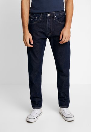 Homme portant un jean droit bleu foncé, une chemise marine et des baskets blanches, debout devant un fond clair uni.