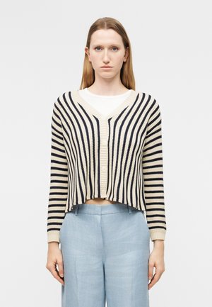 Kobieta ubrana w krótki, beżowo-czarny sweter na guziki w paski, nałożony na białą koszulę, oraz jasnoniebieskie, wysokie spodnie, zwrócona przodem.