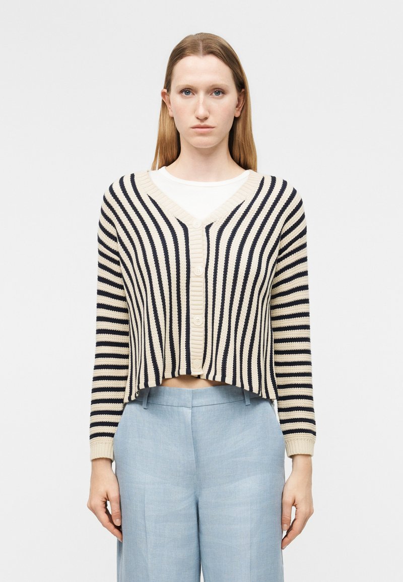 Žena nosi krátky béžovo-čierny pruhovaný zapínací sveter cez bielu košeľu s svetlomodrými nohavicami s vysokým pásom, otočená čelom dopredu.