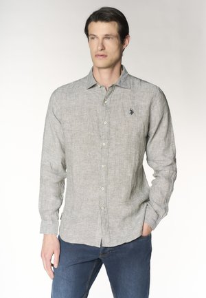 Uomo che indossa una camicia grigio chiaro a maniche lunghe con un emblema scuro sul petto e jeans blu scuro, in piedi con una mano in tasca.