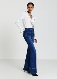 Weißes Blusenshirt mit schwarzem Rüschensaum und langen Ärmeln, kombiniert mit dunkelblauen ausgestellten Jeans mit Taschen und Kontrastnähten. Schwarze High Heels.