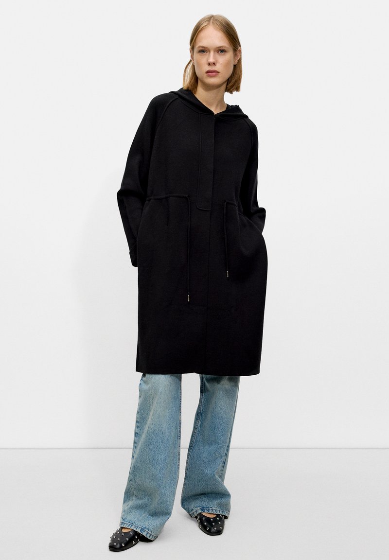 Manteau noir à capuche avec une coupe ample, doté de deux poches avant et de détails à cordon de serrage. Assorti à un jean évasé bleu clair et des talons noirs.