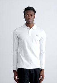 Jauns vīrietis ar kortām cirtainām matiem, tērpies baltā garveida polo krekla un melnās biksēs, stāv pretī vienkāršai gaišai fona.