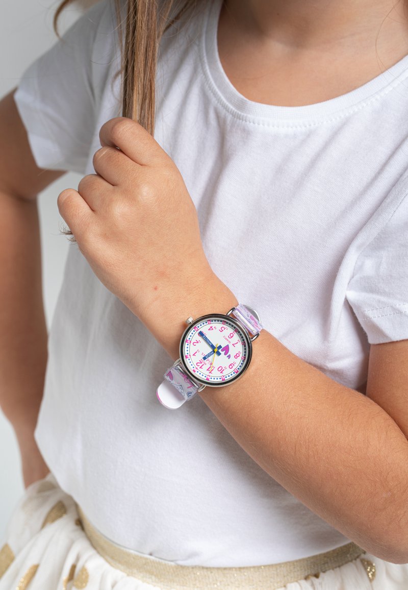 Montre ronde avec un boîtier en argent, cadran blanc, chiffres roses et bracelet en silicone violet à motif. Portée au poignet d'un enfant.