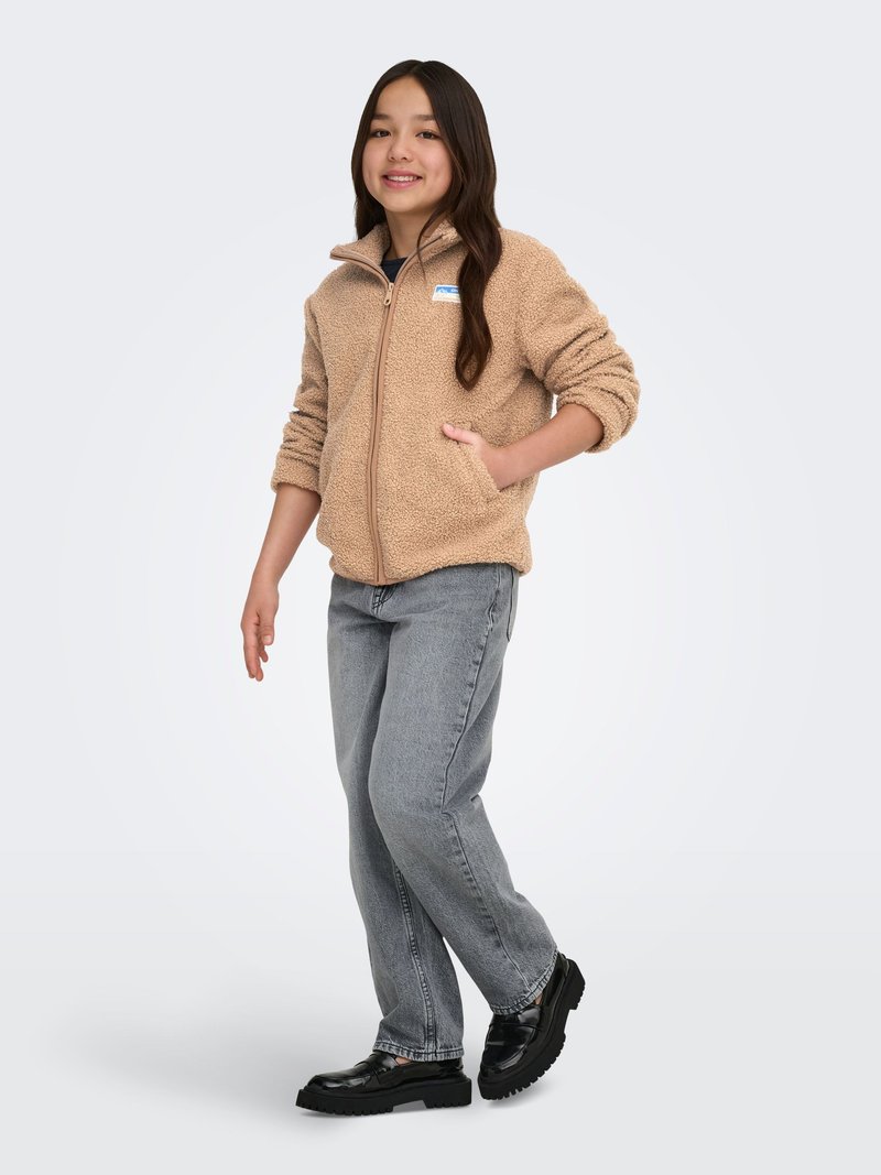 Beige fleecejakke med lynlås, der har en frontlomme og et logo-patch. Båret med grå jeans og sorte platformssko.